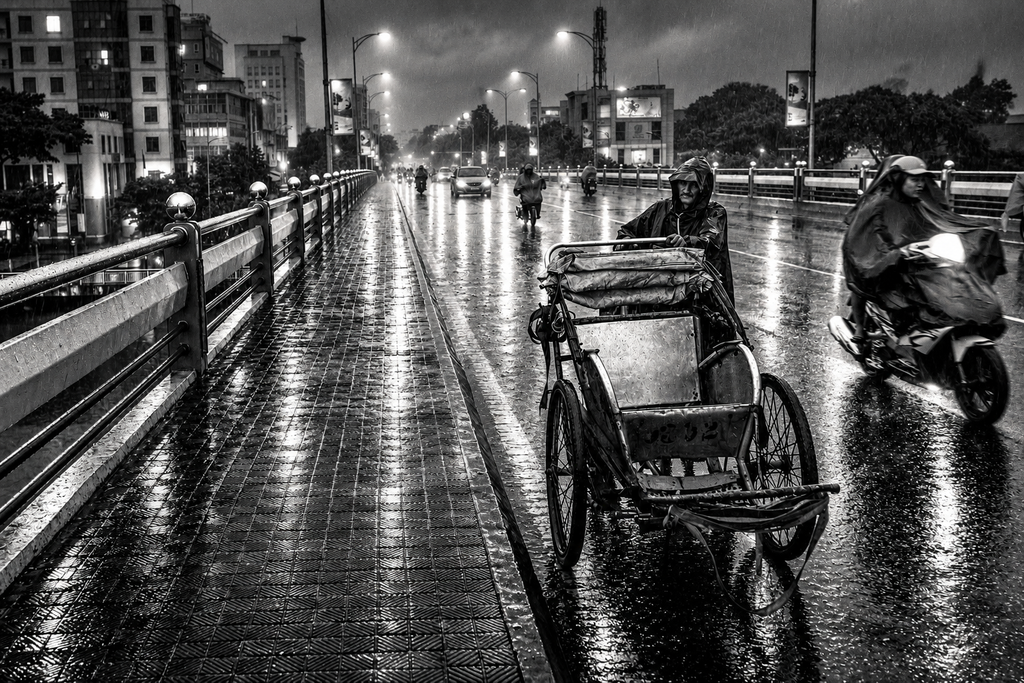 Moody black and white photography of cyclo driver pushing rickshaw across wet bridge on rainy night in Vietnam wall art print